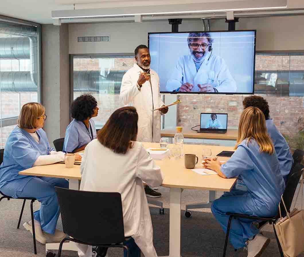 Healthcare professionals in a meeting room, with one presenting and a video call displayed on a screen.