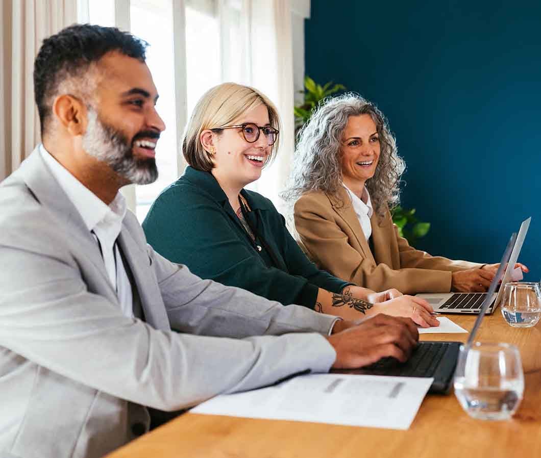 Three people in business attire are working on laptops at a wooden table with documents and glasses of water.