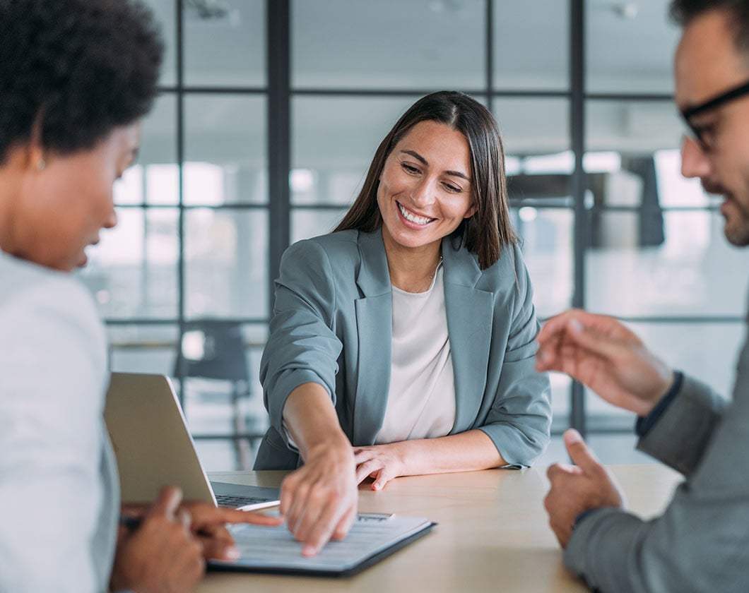 Person pointing at a document during a meeting with colleagues around a table and a laptop.