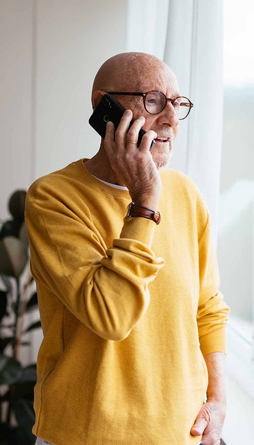 A person wearing a yellow sweater talks on a smartphone by a bright window.
