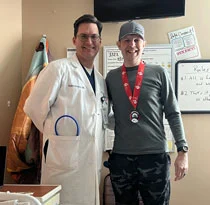 A doctor in a white coat stands beside a person wearing a medal, in an office with notes and posters on the wall.